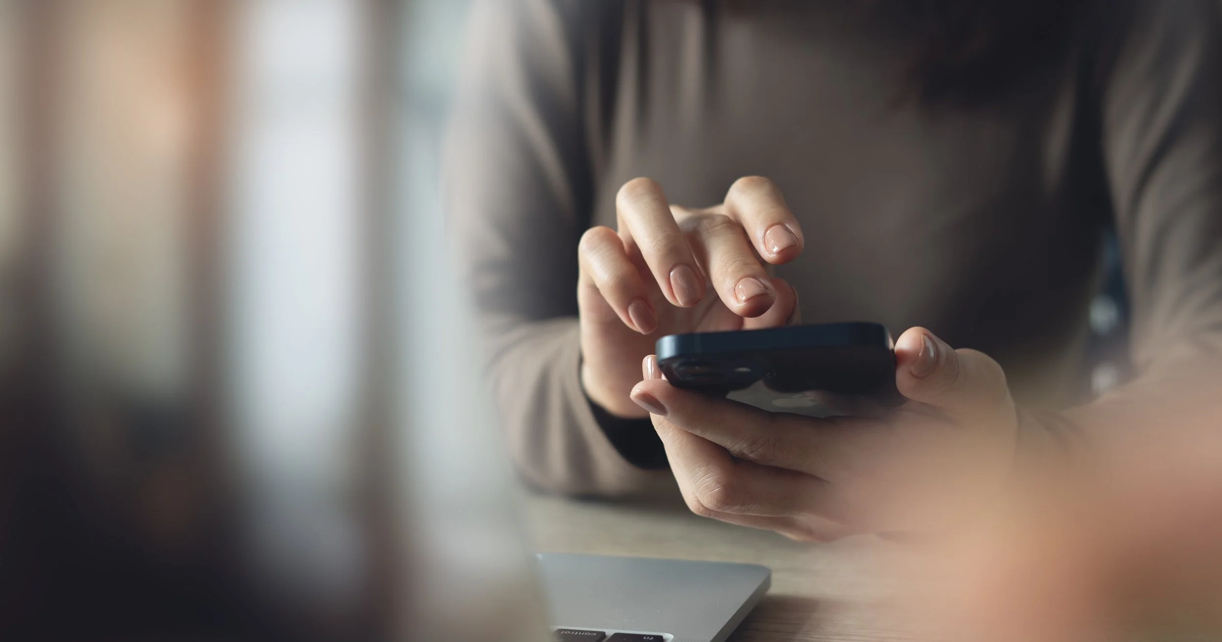 Une personne tient un smartphone noir avec ses deux mains, en train de l'utiliser. La personne porte un vêtement gris ou foncé, et il y a un ordinateur portable sur la table devant elle.