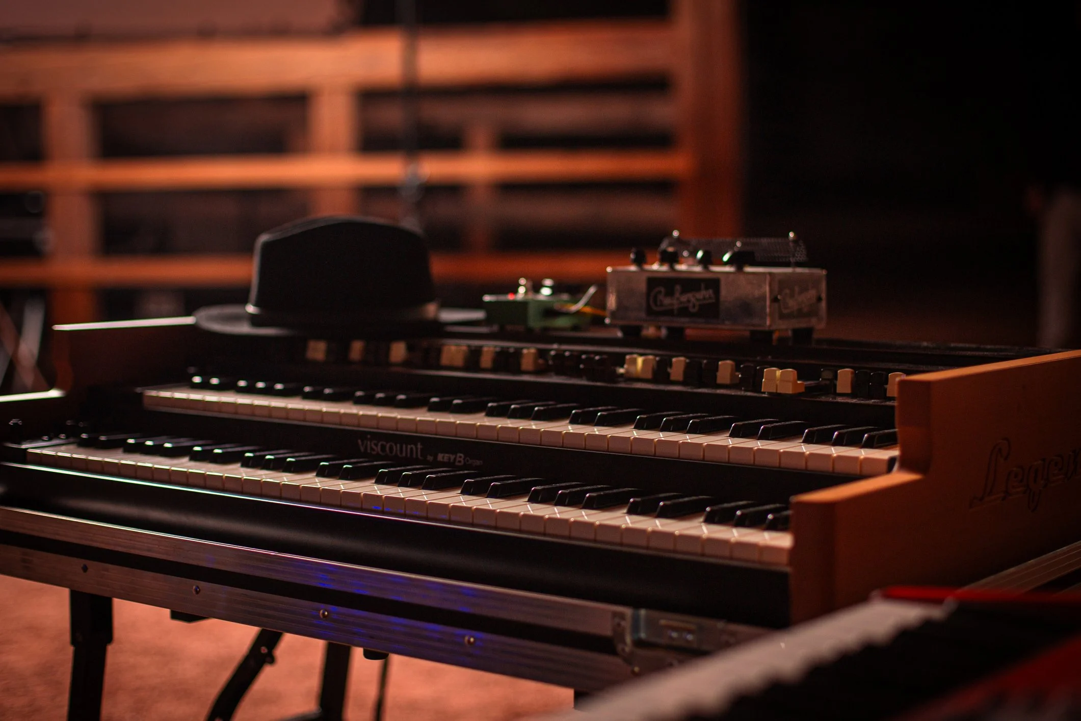 Close-up of a keyboard and a piano, with a hat and a small electronic device on top of the keys, set against a wooden shelf background in a dimly lit room.