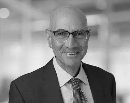 Black and white photo of a man wearing glasses, a suit, and a tie, smiling in an office environment.