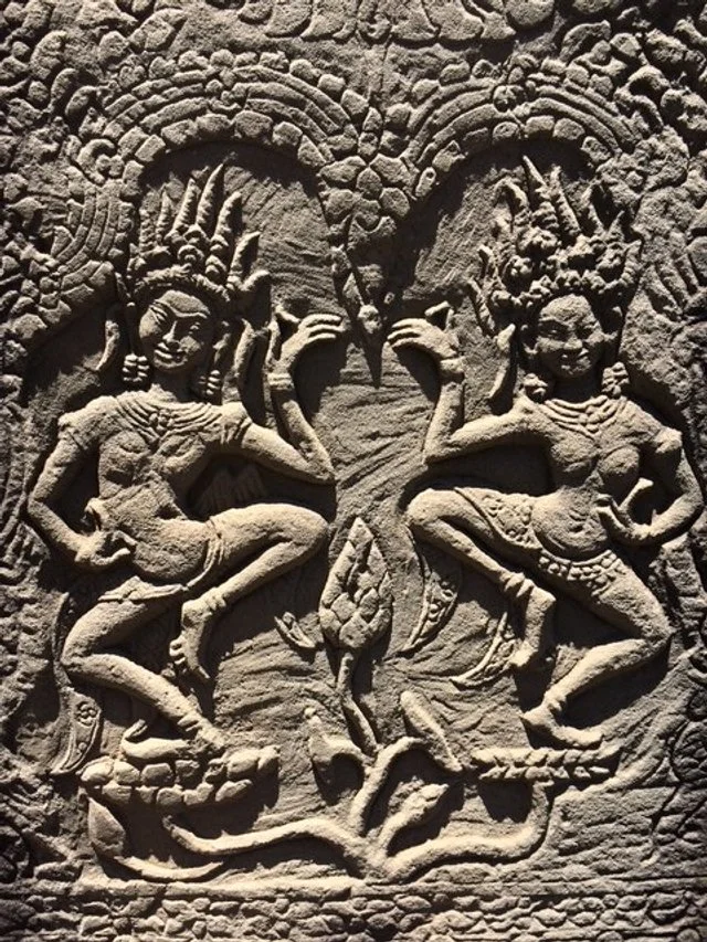 Ancient stone carving depicting two mythological figures with elaborate headdresses, dancing and facing each other with raised hands, surrounded by intricate patterns.