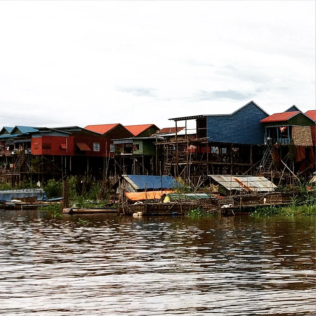 Colorful houses built on stilts along a body of water, with laundry hanging outside some homes and boats floating nearby. Tonle Sap