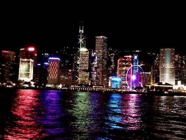 Nighttime Hong Kong skyline and light show from Tsim Sha Tsui Promenade. Illuminated skyscrapers and a lit Ferris wheel reflecting on the water