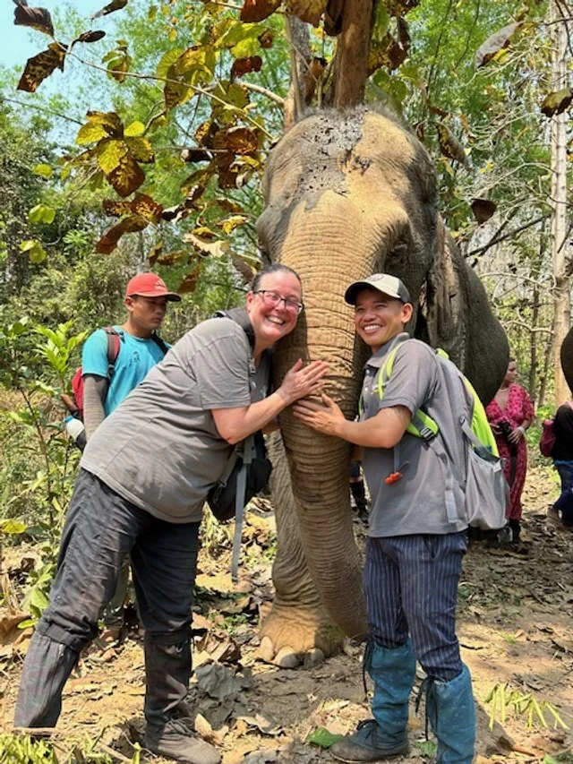 OBT and guide hugging the elephant in a forest setting, with other people visible in the background.
