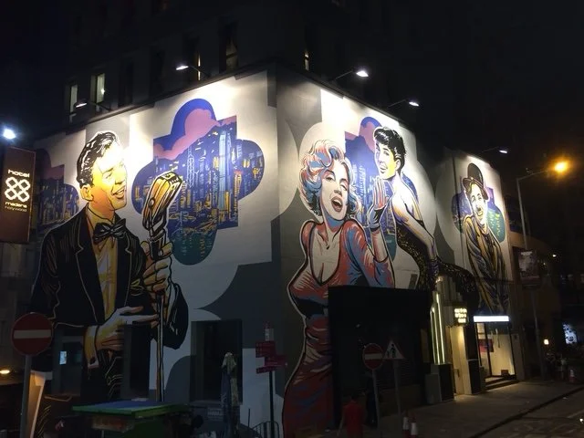Nighttime mural of vintage-style characters on a building wall, including a man in a tuxedo with a microphone, a woman with curly hair singing, and another man with slicked-back hair. Located in Soho area.