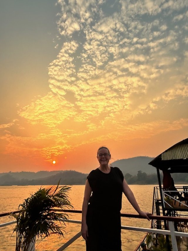 OBT on the boat deck during sunset with a body of water, hills, and a partly cloudy sky in the background. Mekong River Cruise