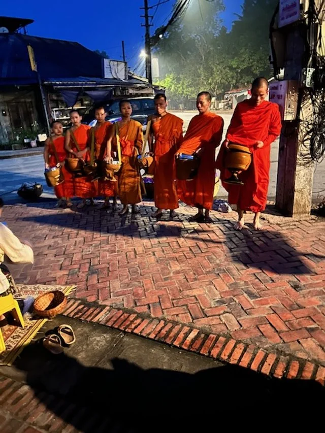 Six Buddhist monks in orange robes walking barefoot along a brick sidewalk at dawn, carrying alms bowls.