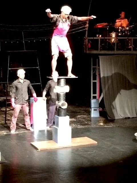 A performer balancing on a stack of black and white blocks on a stage, with arms outstretched. There are other people nearby and a musician playing drums in the background.