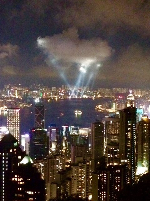Nighttime cityscape of Hong Kong with skyscrapers and Victoria Harbour, with light beams and clouds illuminating the sky.