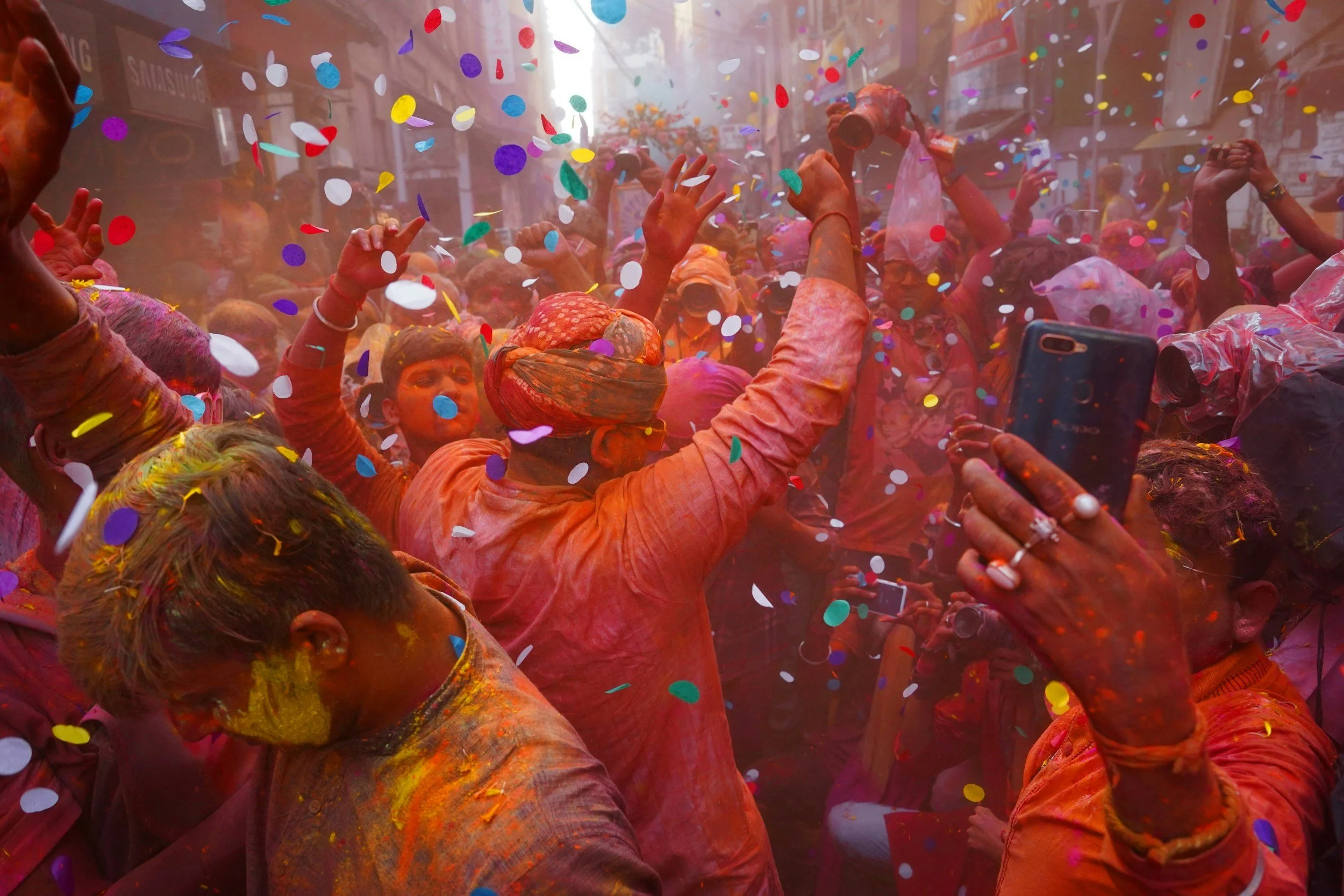 People celebrating Holi festival with colored powders and confetti in a lively street scene.