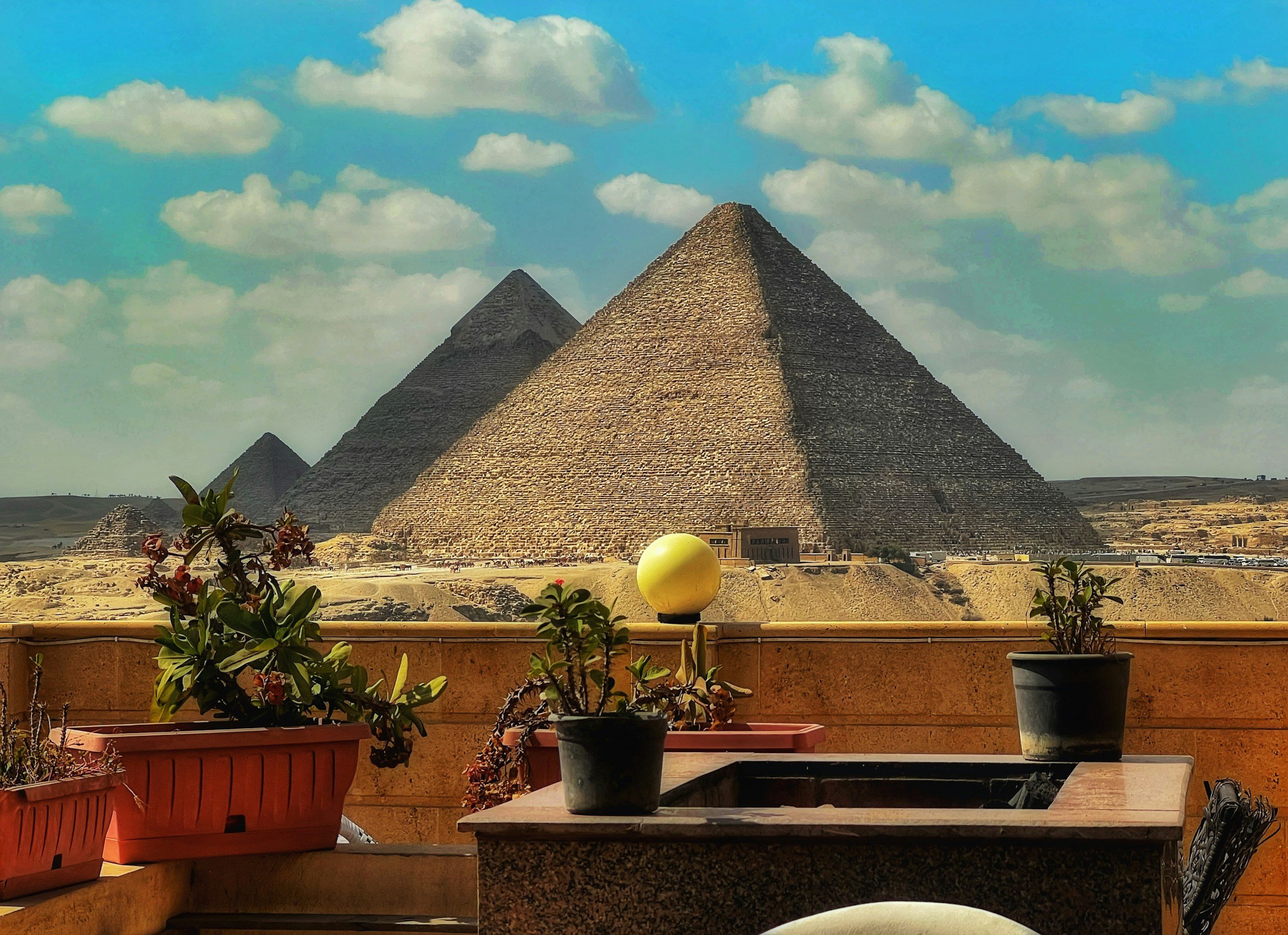 Three pyramids at Giza viewed from a balcony with potted plants in the foreground, under a partly cloudy sky.