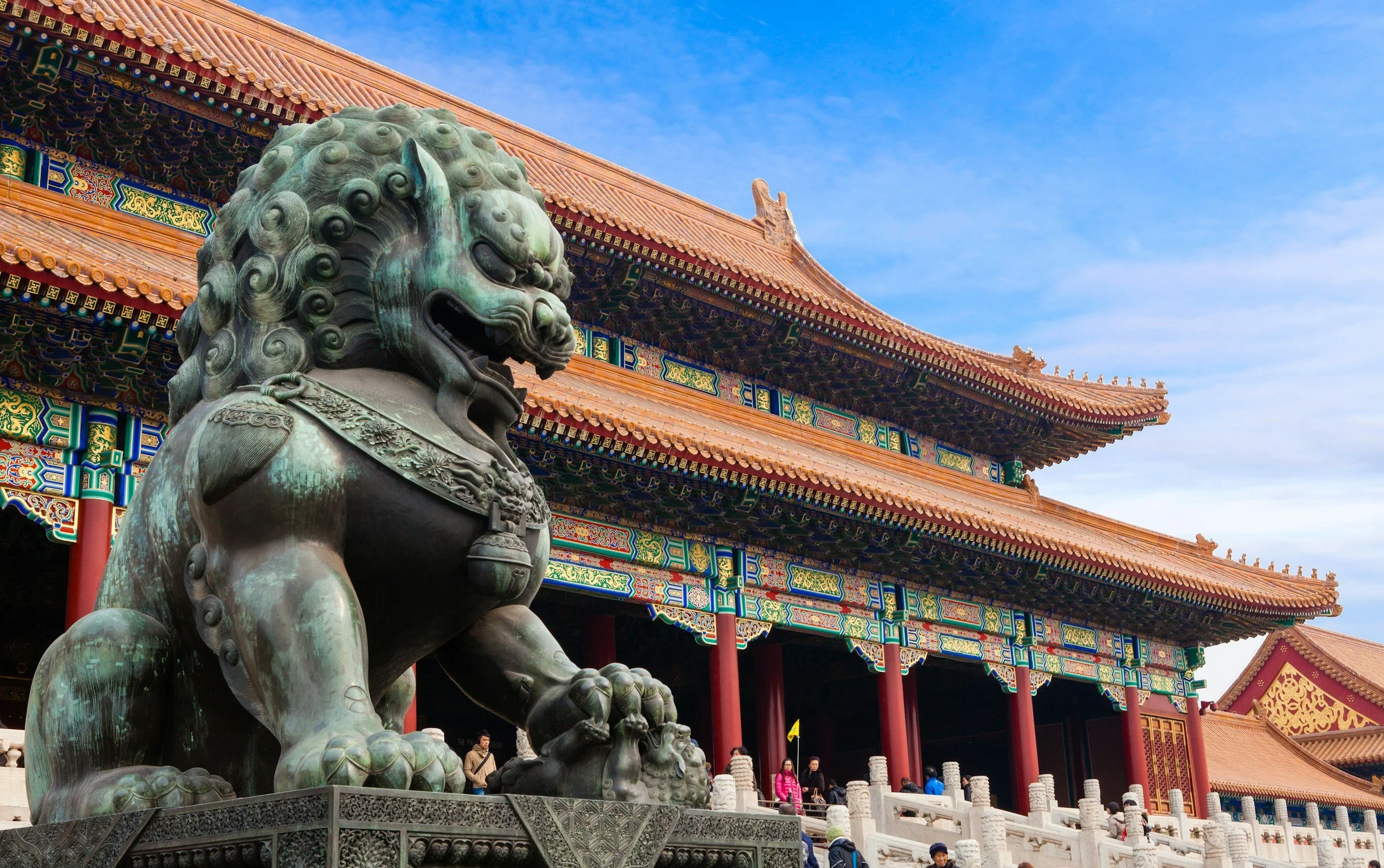 A large bronze guardian lion statue in front of a traditional Chinese temple with ornate, colorful architectural details and orange-tiled roofs under a blue sky.