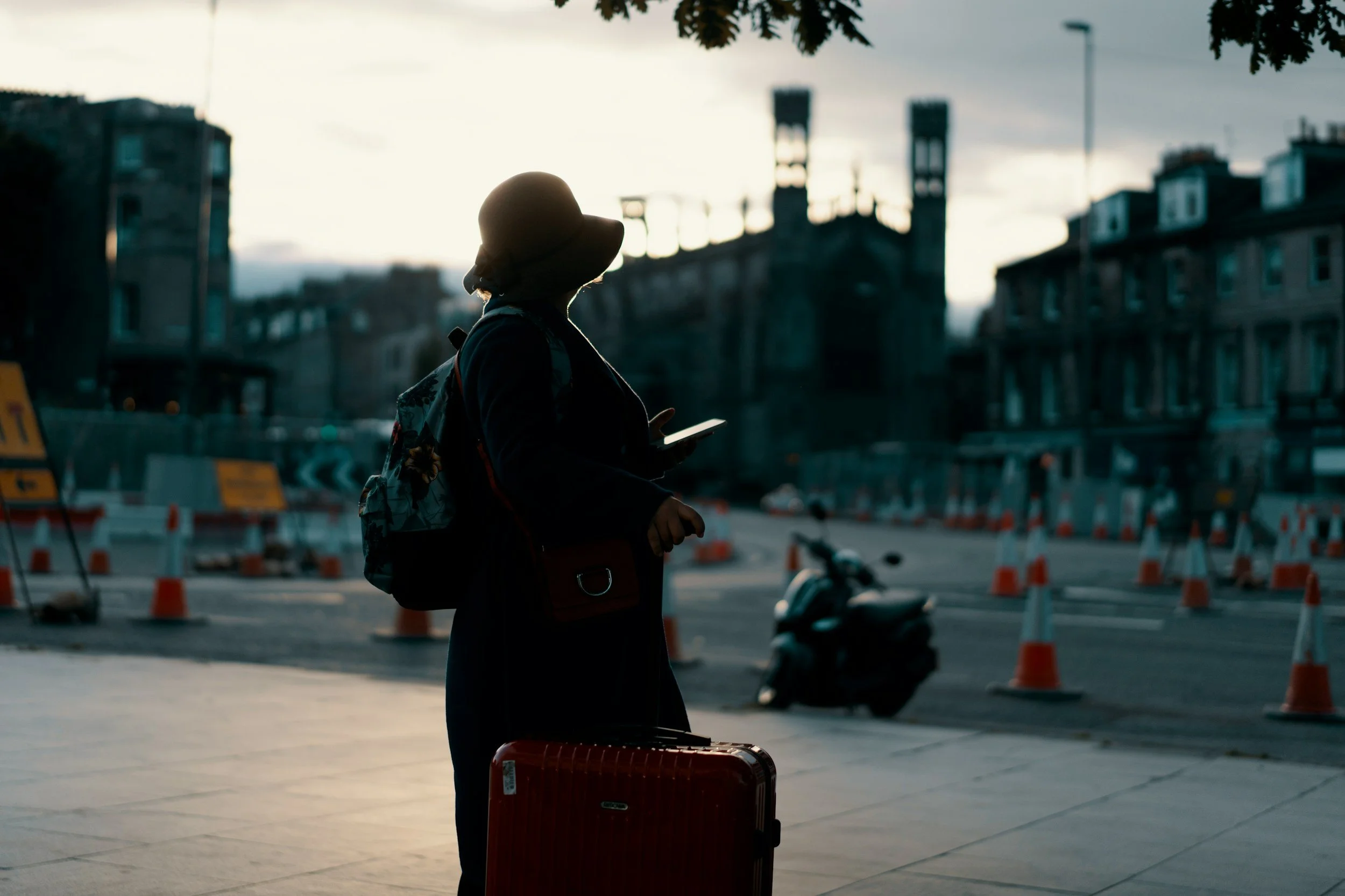 Silhouette of a woman with a hat, backpack, and rolling suitcase standing on a city sidewalk at dusk, looking at her phone. City buildings, traffic cones, and a scooter are in the background.