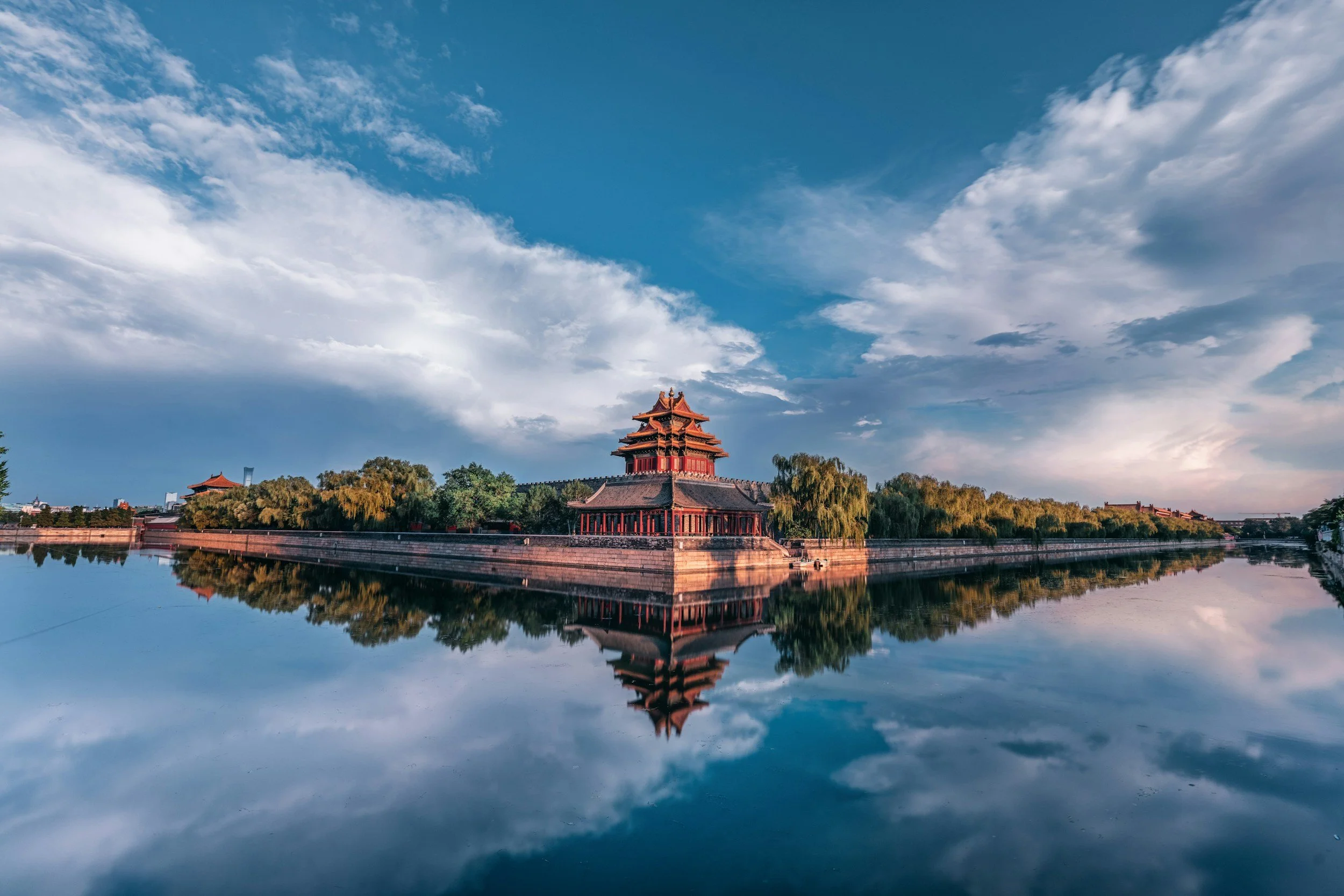 Traditional Chinese pagoda-style building surrounded by trees, reflected in a river under a partly cloudy sky.
