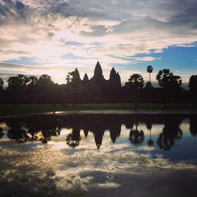 Silhouette of Angkor Wat temple and palm trees reflected on a body of water during sunset or sunrise.