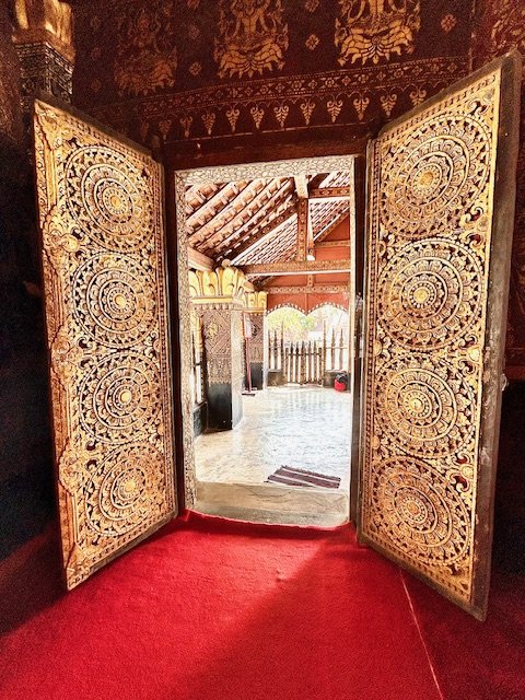 Intricately carved wooden double doors open to an outdoor area with a tiled floor, wooden fence, and a thatched roof structure, framed by decorated interior walls and a red carpeted floor. Wat Xiengthong grounds.