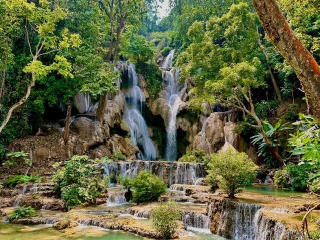 Multiple waterfalls cascading over rocks surrounded by lush green trees and plants in a dense forest. The bigger falls at Kuang Si