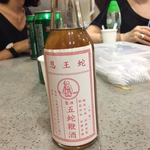 A glass bottle of Chinese snake wine with a pink label, placed on a table with soda cans and people in the background.