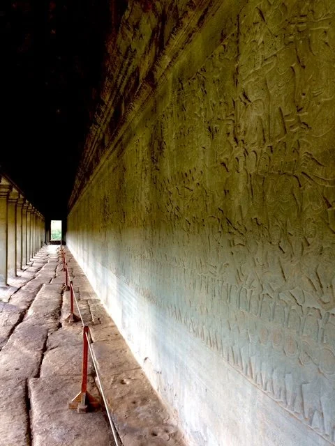 Long stone corridor inside an ancient temple with carved walls on the right, and a row of columns supporting the roof on the left, leading to bright daylight at the end. Inside the main complex with pictographs.
