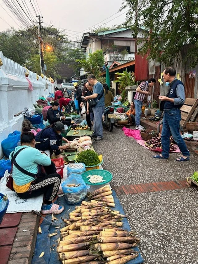 Street market with vendors selling vegetables and root crops, and people shopping along a narrow alleyway.