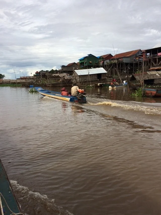 People in a small boat on a river near houses elevated on stilts in a fishing village.