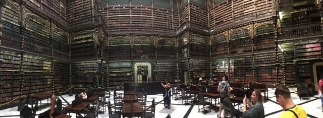 Inside a large, ornate library with tall bookshelves, wooden tables, and chairs, and visitors browsing and studying.