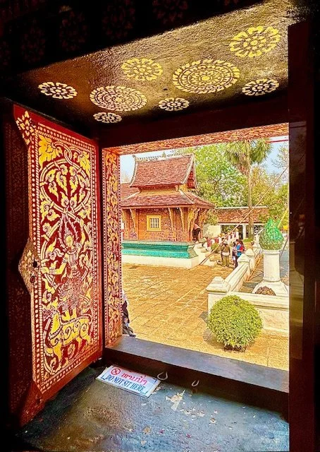 View through an open doorway of a traditional temple courtyard with a small wooden building, sculptures, greenery, and a blue sky, decorated with ornate patterns on the door and ceiling. On Wat Xiengthong grounds.