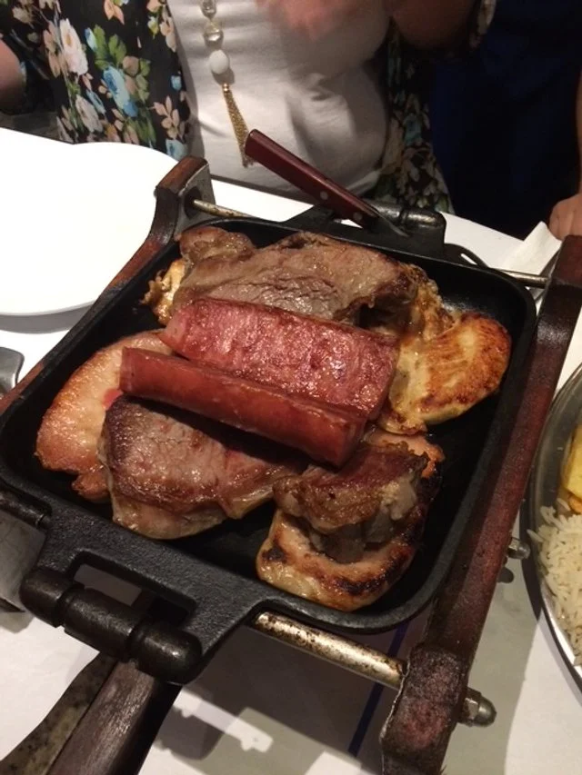 Traditional Brazil casual eats. Cooked steaks, sausage, and potatoes on a sizzling hot grill pan at a dining table.