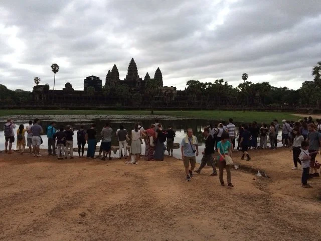 Tourists gather near water with Angkor Wat temple in the background on a cloudy day.