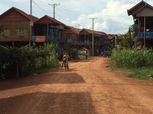 Dirt road in a rural village with wooden houses on stilts on both sides, some with blue railings, and villagers riding bicycles and walking. Power lines run overhead.