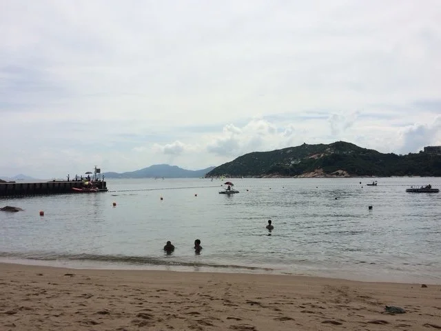 Stanley Beach with calm water, people swimming, boats on the water, a pier on the left, and a hilly island in the background.