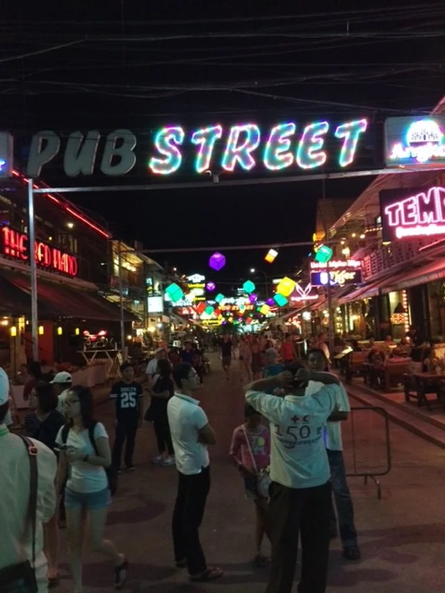Pub street scene at night with neon signs, colorful hanging lanterns, and groups of people walking and socializing. The main strip of bars and restaurants in Siem Reap.