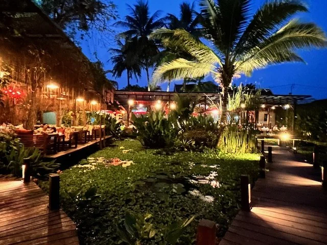 Manda de Laos restaurant at night with wooden walkways, lush greenery, palm trees, and string lights. Absolutely stunning decor.