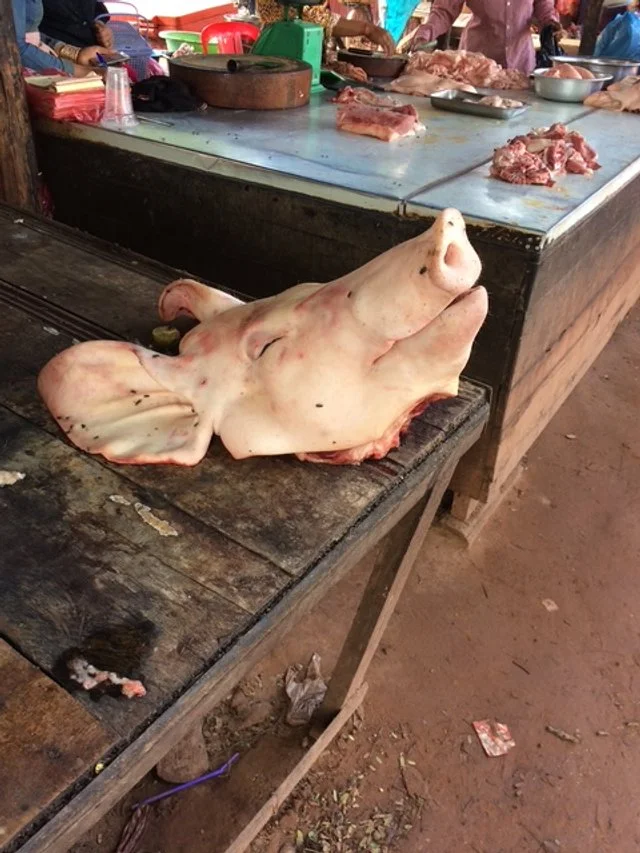 A pig's head and face on a wooden cutting board at a market stall with raw meat and people in the background. Local Market stop.