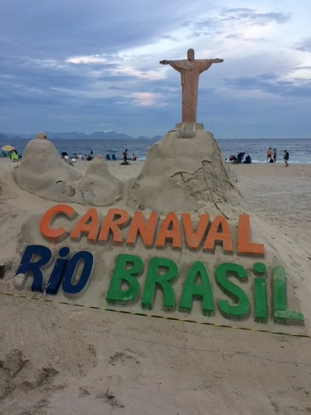 Sandcastle with large colorful letters spelling 'CARNAVAL RIO BRASIL' and a sand sculpture of Christ the Redeemer in the background on a beach with people near the ocean and a cloudy sky.