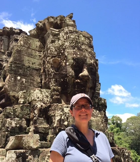 Person smiling and wearing glasses, a cap, and a light-colored shirt, standing in front of an ancient stone sculpture of a face at a historical site, with a blue sky and some clouds in the background.