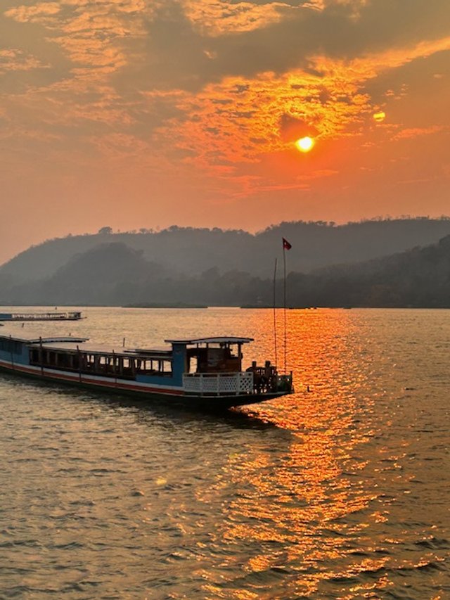 A boat sailing on a river during sunset with a mountain range in the background. Mekong River.