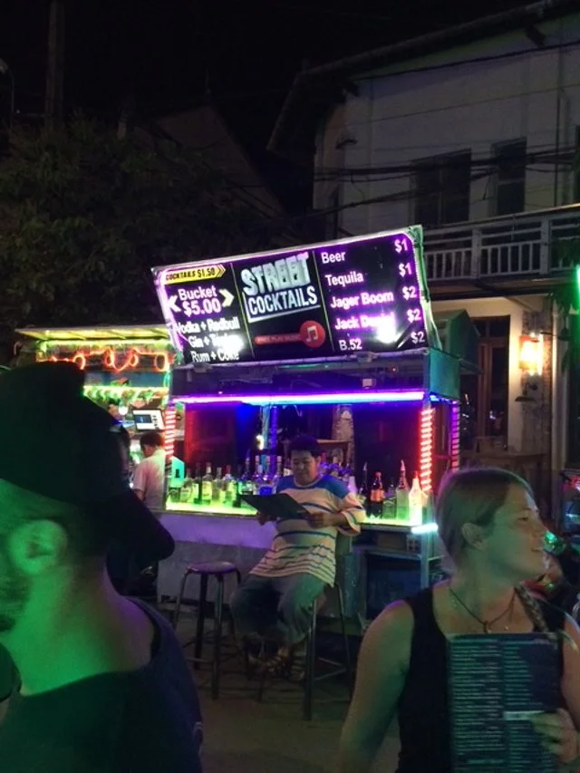 Night scene at an outdoor VW Bus bar with a neon-lit sign advertising street cocktails and drinks. 