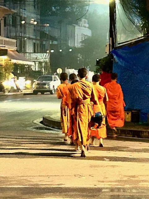 A group of young monks in orange robes walking along a sidewalk in Luang Prabang at dawn.