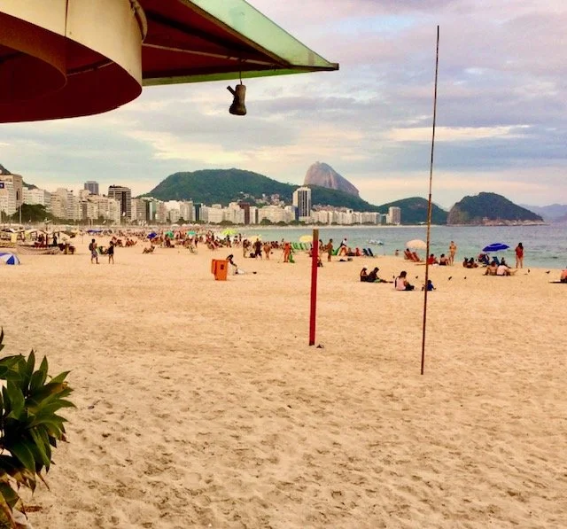 Copacabana Beach scene with people relaxing and playing, city skyline in the background, Sugarloaf Mountain visible in Rio de Janeiro, Brazil, overcast sky.
