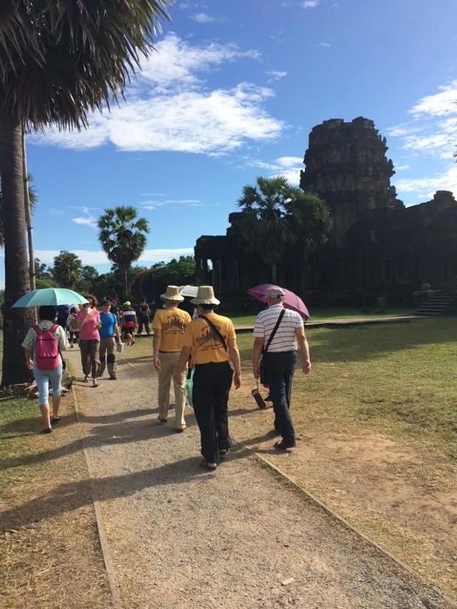 Tourists walking along a path near ancient temple ruins, some holding umbrellas and wearing hats on a sunny day with palm trees and blue sky.