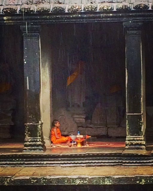 A person sitting cross-legged on a stage or platform, wearing orange clothing, with a small table or tray in front of them holding objects, possibly in a temple or traditional setting with dark stone structures and pillars.