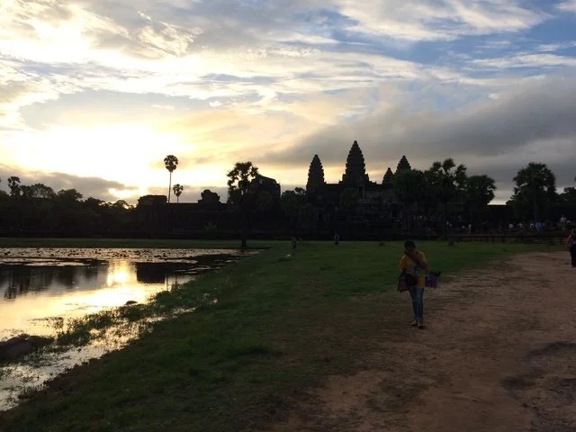 Silhouetted Angkor Wat temple complex at sunset with a person walking on a dirt path in the foreground.