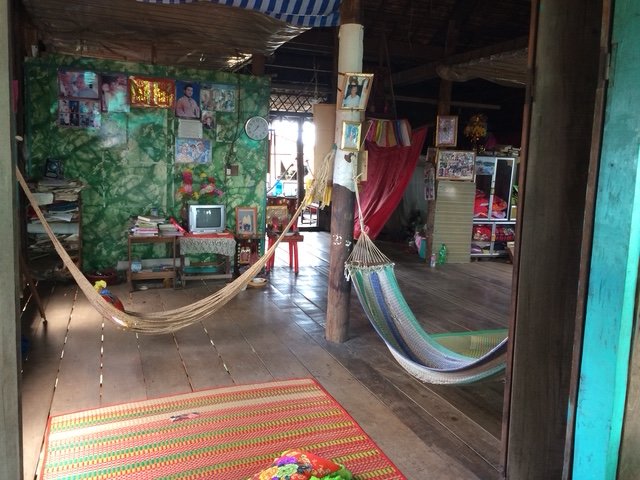 Interior of a rustic home with wooden floors and walls, decorated with colorful fabrics, pictures, and a small TV. Two hammocks hang across the room, and there is a red and orange striped rug on the floor. Various furniture and household items are vi