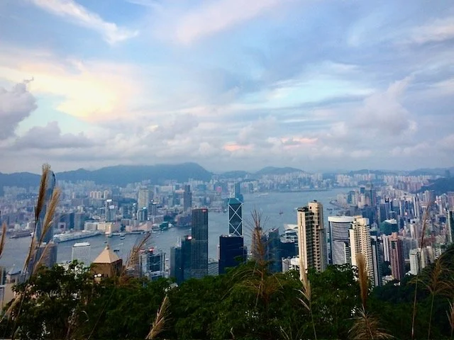 View of Hong Kong skyline with skyscrapers, harbor, and mountains in the background under a partly cloudy sky.