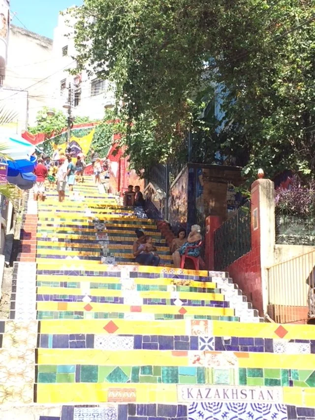Colorful tiled staircase in a hilly area with people sitting and walking, some under umbrellas, leading up to a city scene in Kazahkstan during daytime.