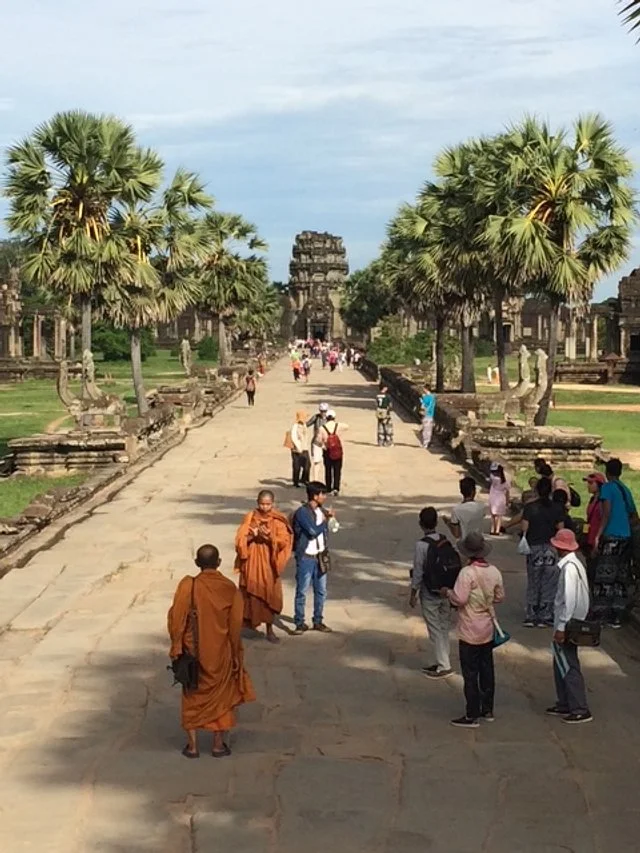 Tourists walking along an ancient stone pathway lined with palm trees, with a temple in the background.