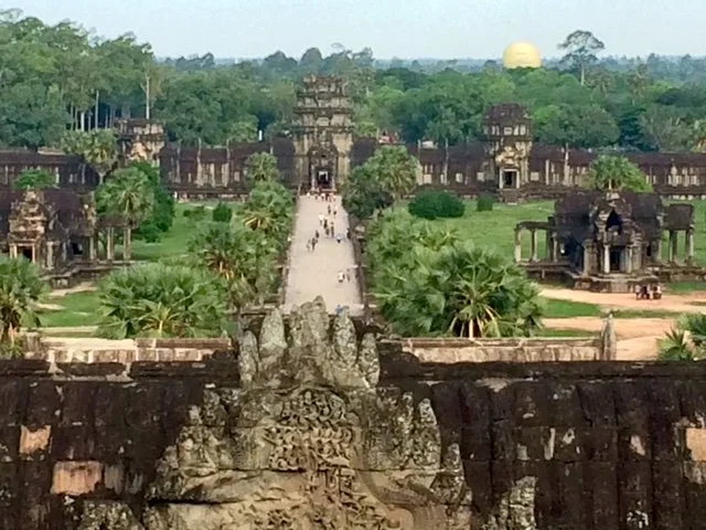 Viewing a historic ancient temple complex surrounded by lush greenery, with a central pathway leading to the main entrance, and the sun setting or rising in the background.