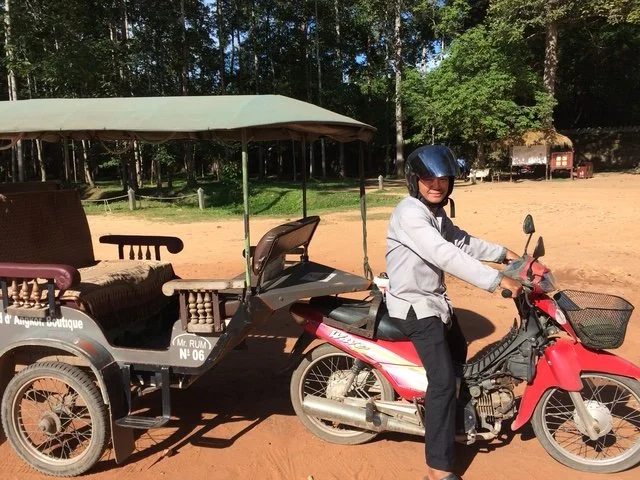Mr Rhum my Angkor Wat driver, pulling a traditional cart on a dirt road, with trees and a few buildings in the background.