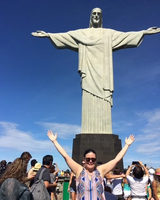 A woman smiling with arms raised in front of the Christ the Redeemer statue in Rio de Janeiro, Brazil, with a clear blue sky and a crowd of tourists.