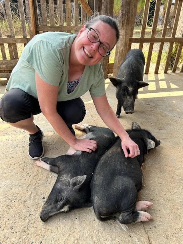 OBT at the Laos Buffalo Dairy, smiling, while petting a group of piglets lying on the dirt. 